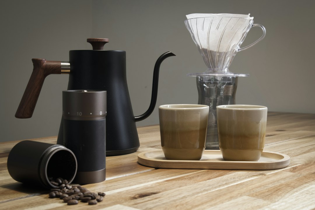 Handmühle mit verstreuten Kaffeebohnen neben einem V60-Handfilter, Schwanenhalskessel und zwei Tassen auf einem Holztisch