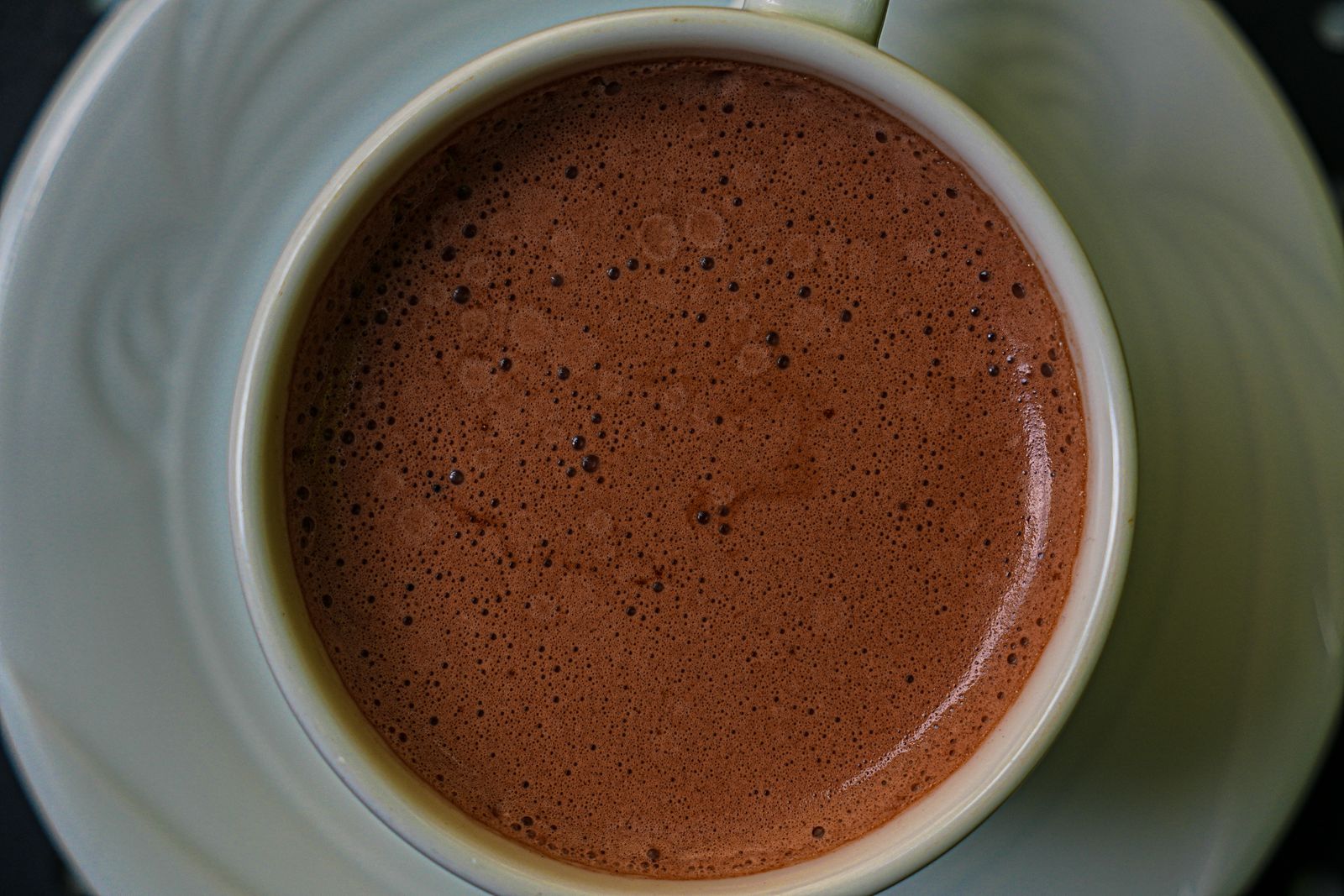 Heiße Schokolade mit feinen Bläschen in einer hellgrünen Porzellantasse auf Untertasse, von oben fotografiert