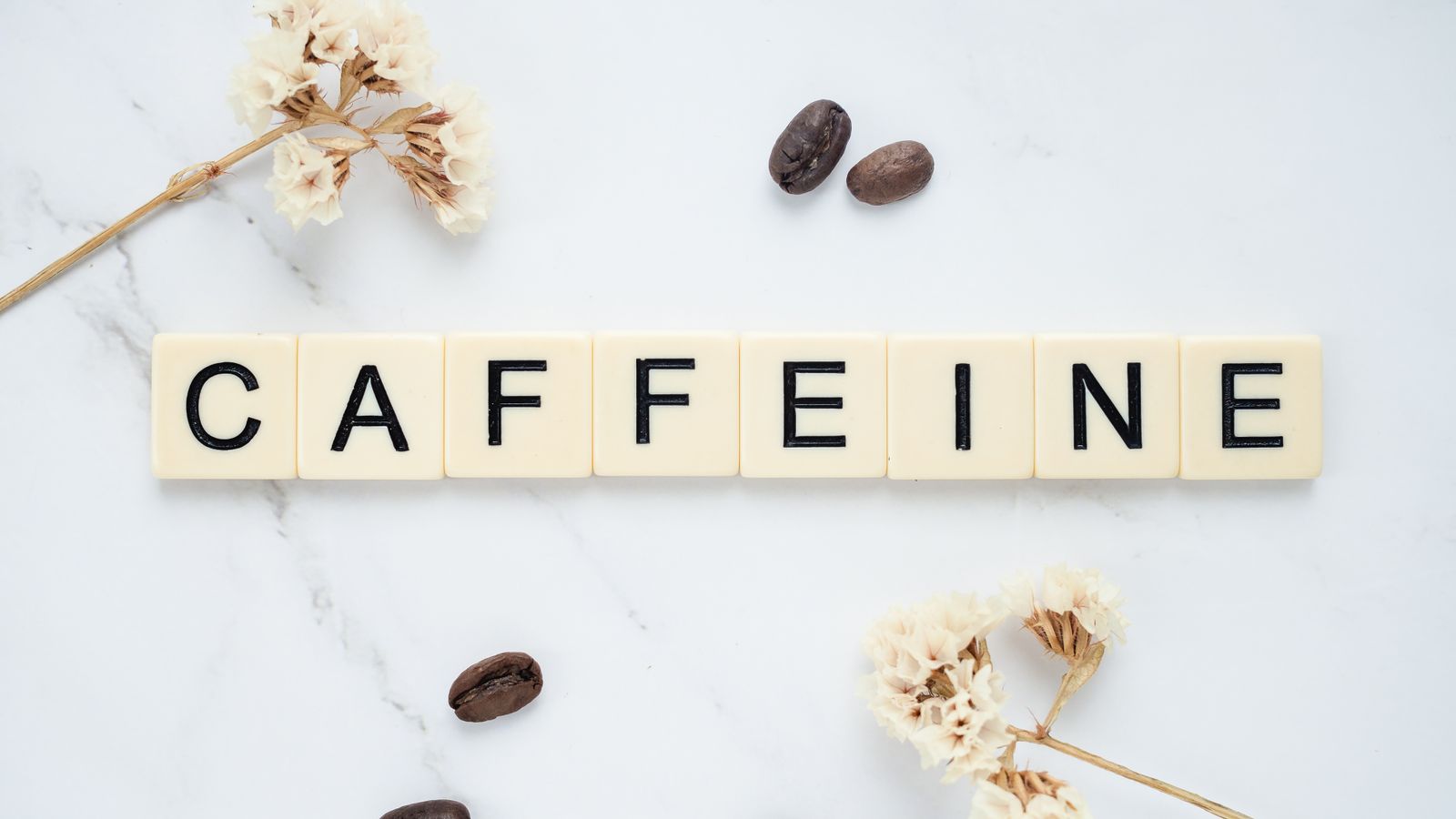 Das Wort „Caffeine“ aus Buchstabensteinen auf weißem Marmor, daneben Kaffeebohnen und getrocknete Blüten