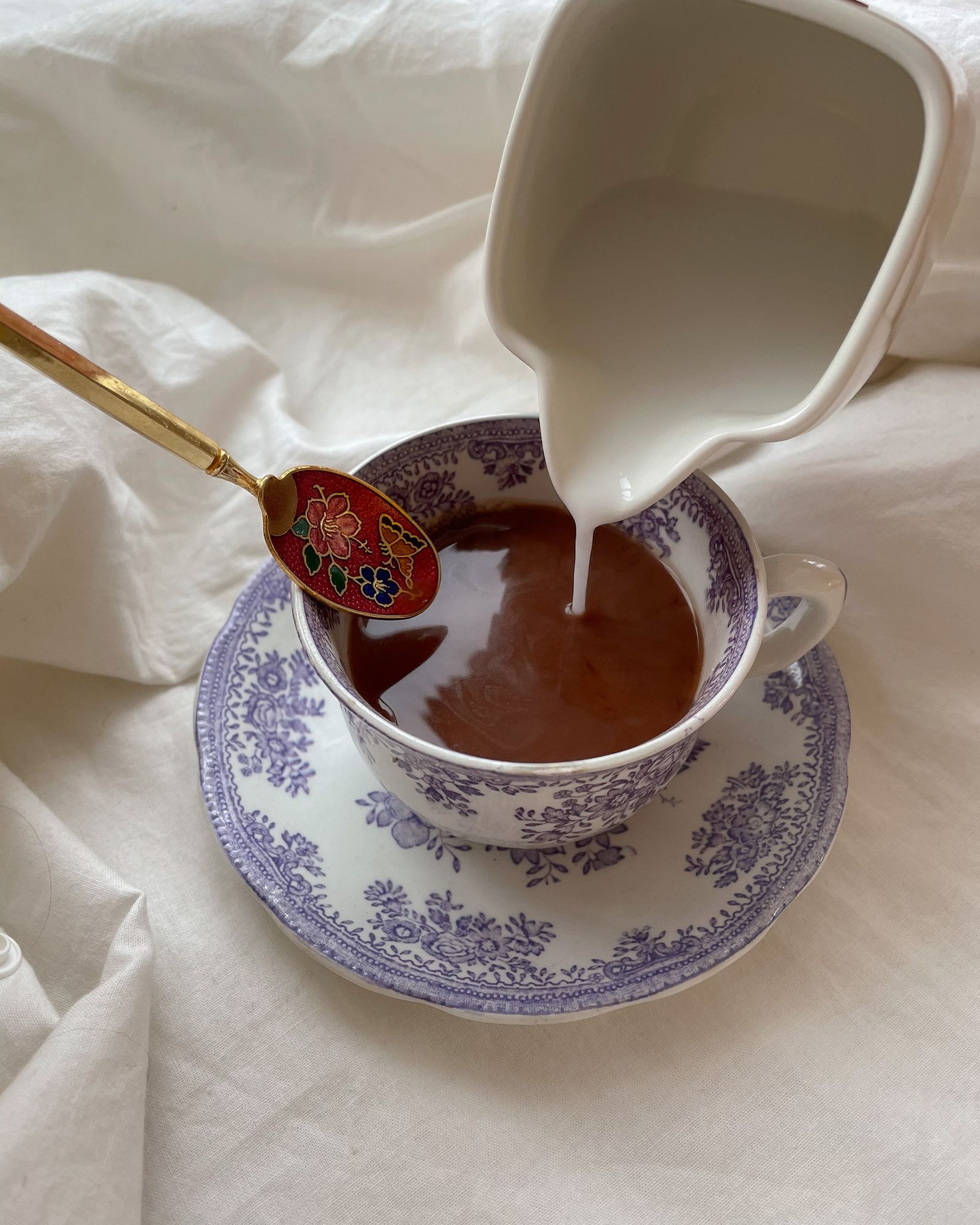 Milch wird aus einem Kännchen in eine Tasse mit Tee gegossen, Porzellan mit lila Blumendekor